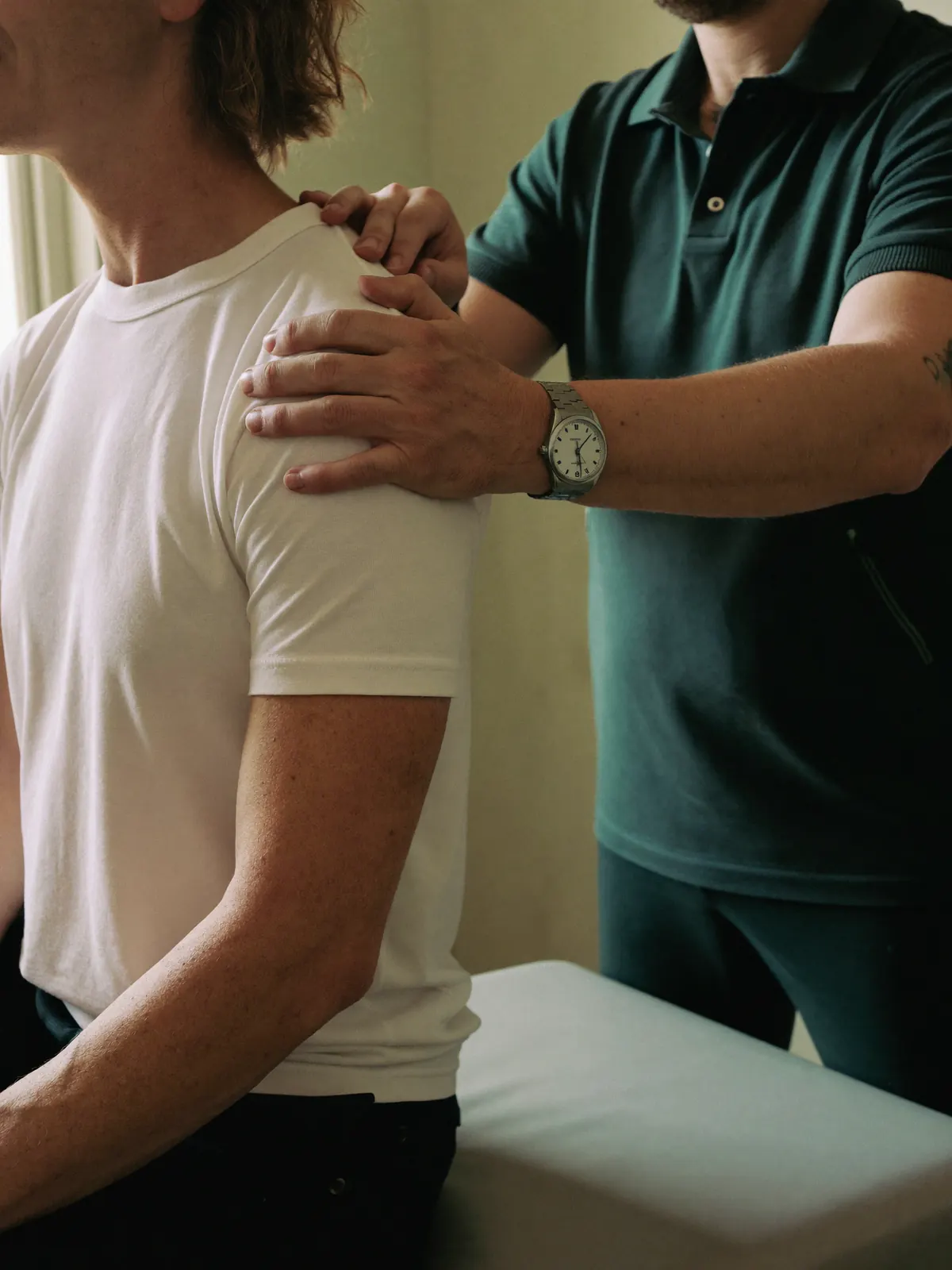 A physio rests both hands on a patient's shoulder to feel how it moves, beside the treatment table.