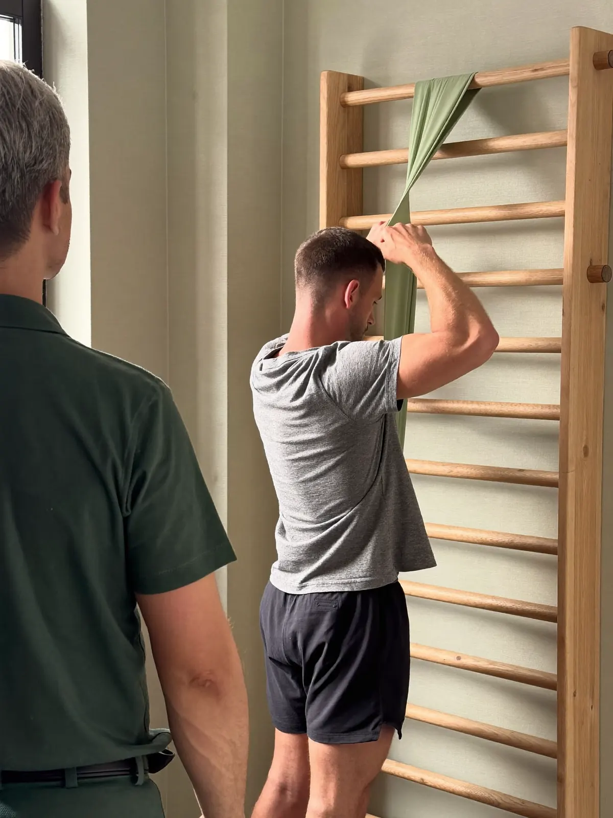 At the wall bars, a man pulls a green resistance band over his head while a physio watches from behind.